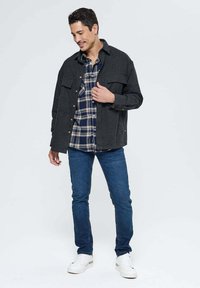 Surchemise en mélange de laine gris, superposée à une chemise à carreaux bleue et blanche, associée à un jean foncé et des baskets blanches. Comprend des poches poitrine.