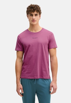 Man with curly hair wearing a purple Marc O'Polo T-shirt and teal drawstring pants, standing against a plain light background.