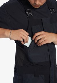 Zwarte overall met een tailored fit, voorzien van verstelbare banden, meerdere zakken en een gladde textuur; een hand plaatst een telefoon in een zak.