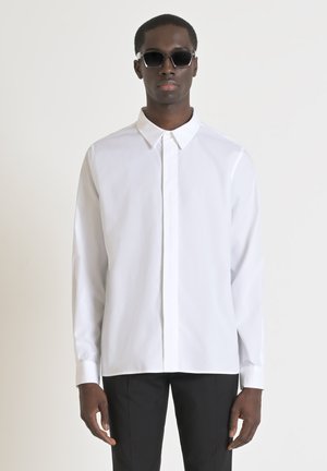 Camisa blanca de manga larga con cuello puntiagudo y tapeta de botones en el frente, combinada con pantalones negros. Tejido suave, sin patrones ni elementos decorativos.