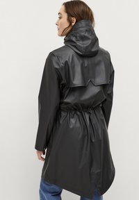Giacca nera impermeabile con cappuccio, vita stretta e orlo obliquo. Materiale liscio e lucido con una piega sul retro per un tocco di dettaglio in più.