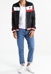 Sort zip-up jakke med røde og hvide striber, kombineret med blå opsmøgede jeans og hvide sneakers. Glat materiale og tætsiddende design.