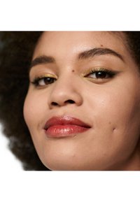 Gros plan sur un visage aux cheveux bruns bouclés. Les traits incluent un rouge à lèvres bronze, un fard à paupières doré et une texture de peau lisse avec quelques imperfections mineures.