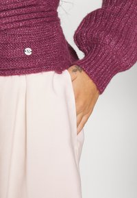 Main avec un petit tatouage reposant dans la poche d'un pantalon rose clair, portant un pull en maille violette texturée avec une petite étiquette métallique avec logo.