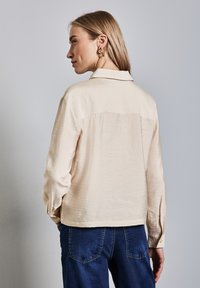 Blusa beige a maniche lunghe con colletto, stoffa textured e polsini con bottoni, abbinata a jeans in denim blu.