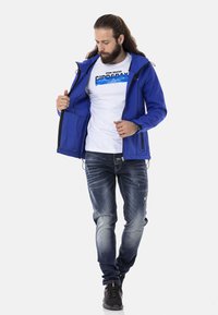 Blaues Softshell-Jacke mit einem Reißverschluss, zwei Seitentaschen, über einem weißen Grafik-T-Shirt getragen, kombiniert mit verwaschenen blauen Jeans und schwarzen Turnschuhen.