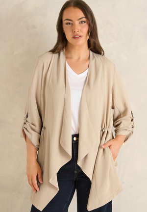 Femme aux longs cheveux bruns portant un cardigan beige ouvert sur un chemisier blanc et un jean foncé, debout devant un fond clair et texturé.