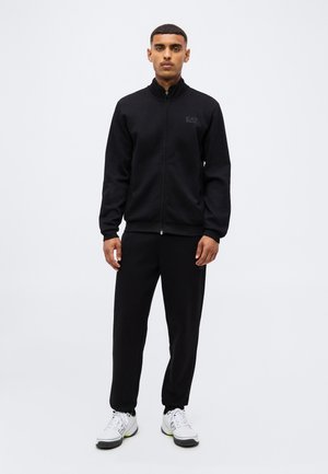 Homme debout, de face, portant une veste noire à fermeture éclair, un pantalon noir et des baskets blanches sur un fond clair uni.
