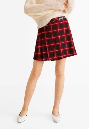 Mujer con falda plisada de cuadros rojos y negros combinada con un suéter oversize color crema y tacones blancos de punta afilada.