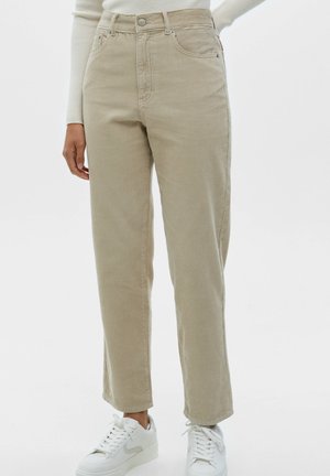 Personne portant un pantalon beige en velours côtelé taille haute avec poches avant et des baskets blanches sur un fond blanc uni.