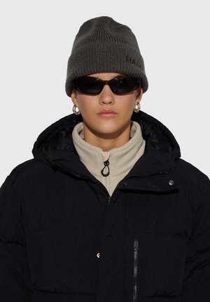 Schwarze Steppjacke mit hohem Kragen, getragen über einem beigen Fleece-Oberteil. Graue Strickmütze und dunkle Sonnenbrille vervollständigen das Outfit.
