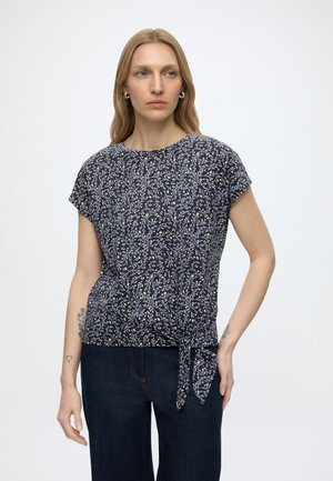 Kobieta o długich blond włosach, ubrana w granatową kwiatową bluzkę z krótkim rękawem zawiązaną w talii oraz ciemnoniebieskie dżinsy, stojąca na jednolitym tle.