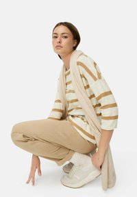 Gestreiftes Langarmhemd in Creme und Beige, beige Hose, beige Sneaker mit weißen Sohlen, leichter Schal über der Schulter drapiert, hockende Pose.