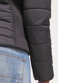 Svart vadderad jacka med ribbad textur längs nedre kanten, tryckknappsstängning och en rundad skärning vid ärmsluten, kombinerad med ljusblå denim.