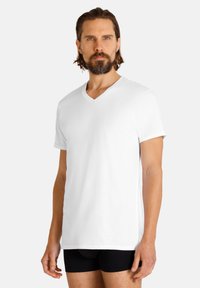 T-shirt bianco a scollo a V realizzato in cotone morbido, maniche corte, con una vestibilità rilassata. Indossato con pantaloni corti neri, caratterizzati da una texture liscia.