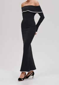 Vrouw in een fitted off-shoulder zwart jurkje met witte randen en een uitlopende zoom, gecombineerd met zwarte hoge hakken met uitsnijdingen.