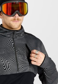 Zwart-witte zip-up jack met golvende lijnpatroon, zwarte onderkant en opstaande kraag; model draagt oversized skibril.