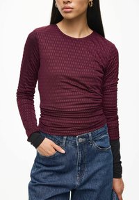 Bordeaux lange mouwen top met een textuurpatroon, fitted ontwerp en zwarte laagdetails bij de manchetten. Gecombineerd met hoge taille denim jeans.