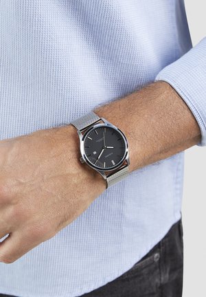 Black dial watch with silver mesh band, featuring a round case, minimalist design, and date function. Worn on a wrist against a light blue shirt.