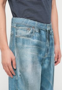 Άτομο που φοράει φθαρμένα ανοιχτό μπλε τζιν και μπλε σκούρο και λευκή ριγέ μπλούζα, με τα χέρια χαλαρά στο πλάι.