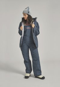 Navy blue winter jacket with gray accents, paired with a jumpsuit featuring a zip pocket. Model wears a gray beanie and ski socks.