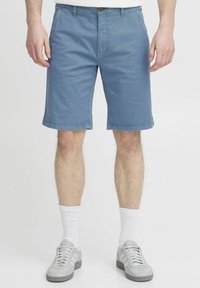 Shorts bleu clair en coton avec un design à plat, passants de ceinture et fermeture à bouton. Portés avec des baskets grises et des chaussettes blanches.