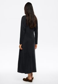 Largo vestido negro hecho de tejido suave, con mangas largas, un corte holgado y un dobladillo recto. Llevado con zapatos marrones sin cordones.