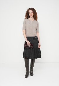 Γυναίκα με σγουρά μαλλιά που φοράει μπεζ κουμπωτή ζακέτα, μαύρη πλισέ φούστα με αγκράφες, μπότες έως το γόνατο, κρατώντας ένα σκούρο κόκκινο τσαντάκι.