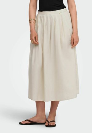 Vrouw met een crème kleurige geplooide midirok, zwarte top, gouden armbanden en zwarte sandalen, staand tegen een effen achtergrond.