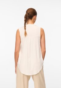 Blusa senza maniche color crema con orlo rotondo, pieghe sottili sul retro e una texture liscia. Abbinata a pantaloni beige chiaro.