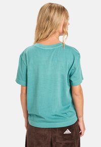 T-shirt in cotone turchese con maniche corte e vestibilità rilassata. Dettagli notevoli includono un collo rotondo e orlo cucito. Indossato con pantaloni in corduroy marroni.