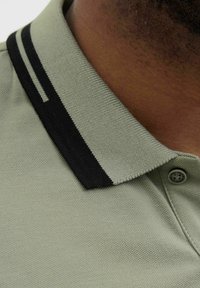 Polo majica svijetlozelene boje, teksturirane tkanine, s crnim ovratnikom koji ima dvije bijele pruge. Uključuje detalj s gumbom na ovratniku.