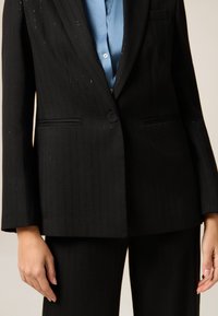 Czarna marynarka w prążki, z długimi rękawami, dwoma przednimi kieszeniami i pojedynczym zapięciem na guzik. Noszona na jasnoniebieskiej koszuli. Dopasowany krój.