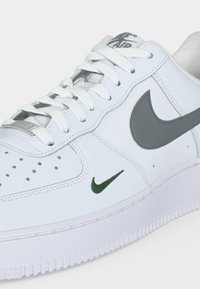 Λευκό αθλητικό παπούτσι Nike Air Force 1 με λευκά κορδόνια, γκρι σύμβολο Nike, μικρό πράσινο σύμβολο κοντά στη σόλα και τρυπήματα στο μπροστινό μέρος.
