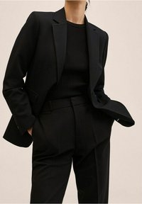 Henkilö, joka on pukeutunut mustaan räätälöityyn pukuun, jolla on kankainen musta paita, kädet rennosti taskuissa, yksinkertaista vaaleanbeigeä taustaa vasten.