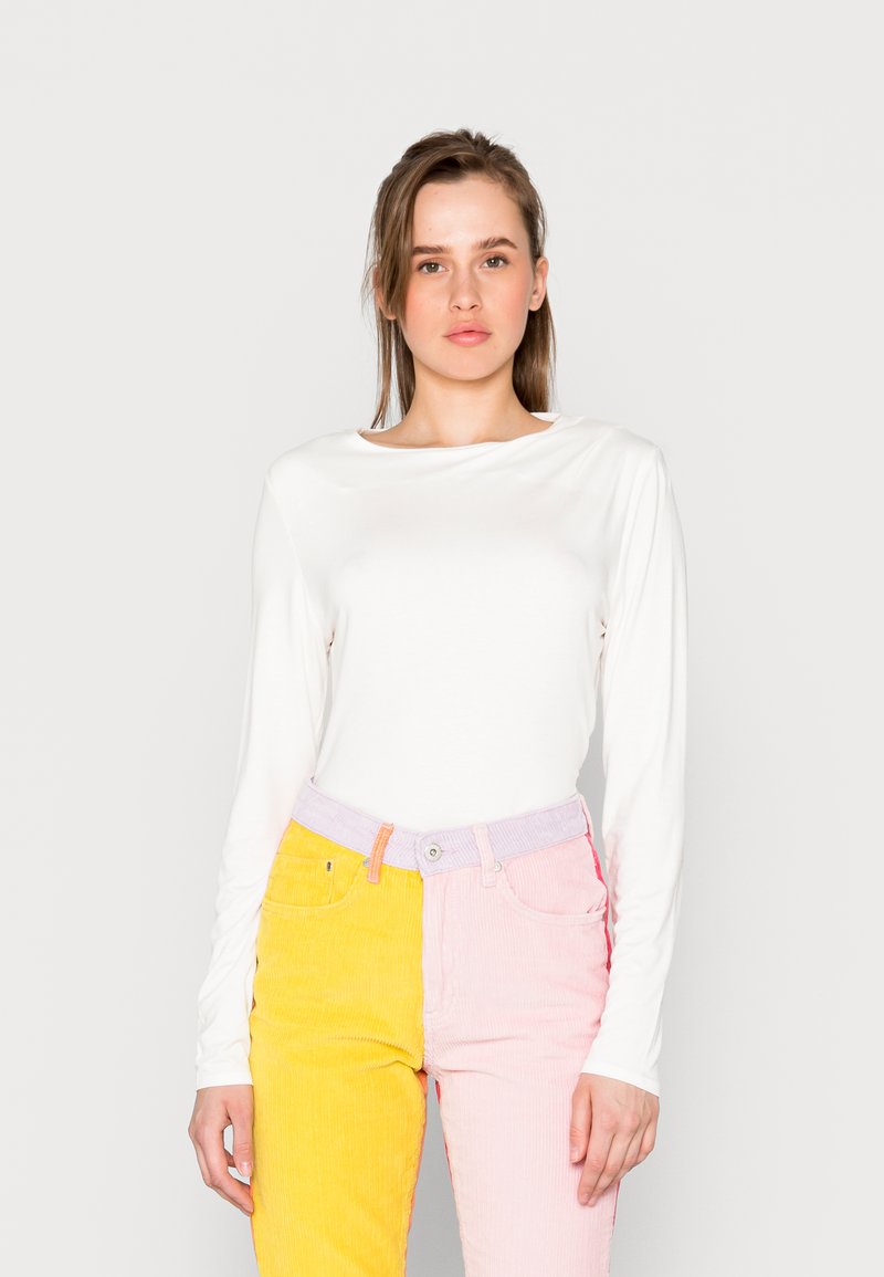 Camisa branca de manga longa combinada com calças de veludo cotelê divididas verticalmente—amarelo do lado esquerdo e rosa do lado direito, apresentando uma faixa na cintura de um roxo claro.