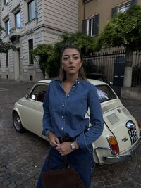 Gaiši zila džinsu krekls ar pogām apkārt apkaklei, kombinēts ar tumši zilām džinsa biksēm. Brūnas zamšādas rokassoma un nerūsējošā tērauda pulkstenis. Fona redzama vintažas stila krēmkrāsas automašīna.