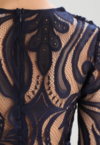 Marineblaue Spitzenbluse mit kunstvollen Blumenmustern, die ein durchsichtiges Design, klare Formen und einen Reißverschluss auf der Rückseite aufweist.