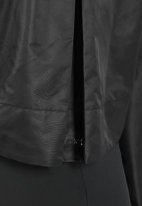 Čierna bunda vyrobená z hladkej tkaniny, s predným zipsom a rozdeleným dizajnom na spodnej časti. Minimalistický štýl bez viditeľných vzorov.
