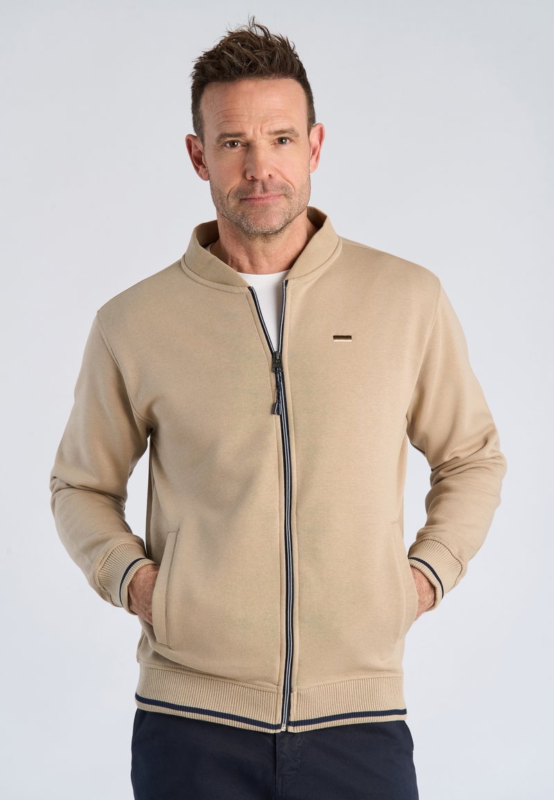 Uomo con indosso una giacca beige con zip, polsini e orlo a coste, in piedi con le mani nelle tasche davanti a uno sfondo chiaro e uniforme.