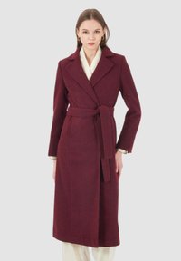 Cappotto in misto lana borgogna con vita cintata, rever a punta, maniche lunghe e lunghezza media. Tessuto liscio e vestibilità su misura.