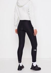 Leggings negros con una cintura alta, textura elegante y detalle de logo blanco en la parte inferior de la pierna. Combinados con un hoodie gris claro y zapatillas negras.