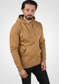 Brun hættebluse lavet af blødt stof med en lomme foran, snor i hætten og ribbede manchetter, parret med sorte jeans. Glat tekstur.