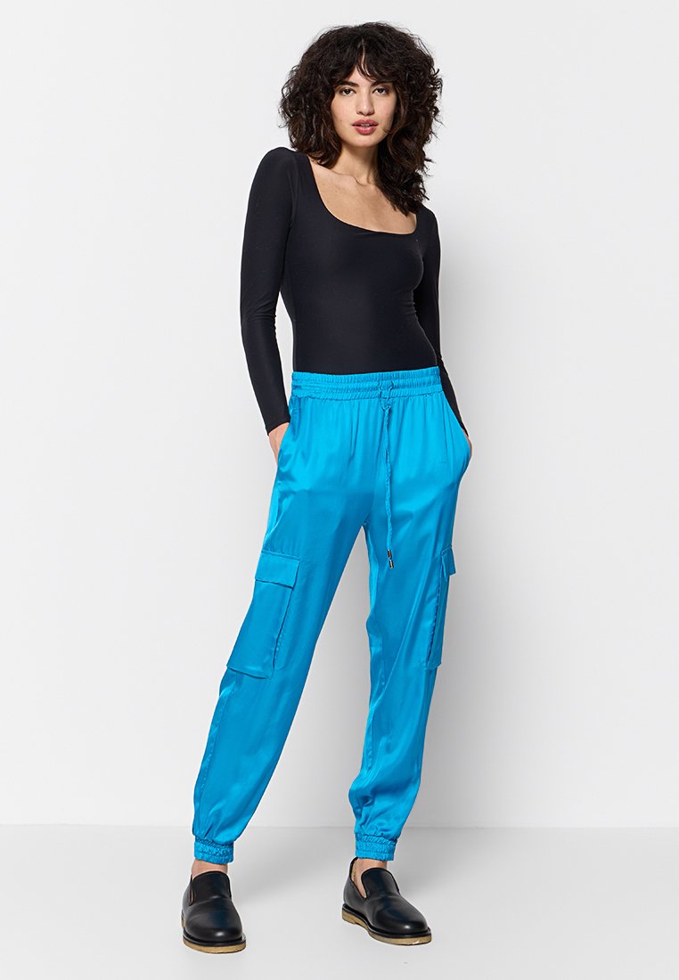 Femme aux cheveux bouclés portant un haut à manches longues noir, un pantalon cargo bleu vif et des chaussures noires à enfiler, se tenant les mains dans les poches.