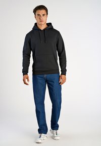 Svart hoodie med ficka fram, kombinerad med blå jeans och vita sportskor. Slät tygtextur, avslappnad passform.