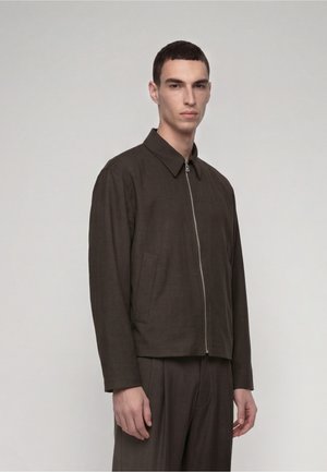 Man wearing dark brown zip-up jacket with collar and matching trousers, standing against a plain light gray background.
