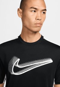 Černé sportovní tričko s kulatým výstřihem, na kterém je velké bílé tečkované logo Nike přes prsa. Krátké stříbrné detaily na ramenech.