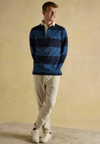 Junger Mann lächelt, trägt ein blau gestreiftes Rugbyshirt, beige Cordhosen und weiße Sneaker und steht vor einer einfarbigen beigen Wand.