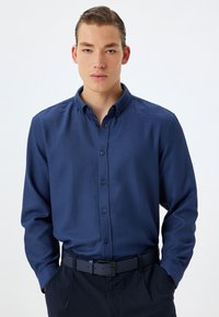 Camisa de botones de manga larga azul marino con una tela texturizada, cuello abotonado y botones a juego, metida dentro de pantalones oscuros con un cinturón.