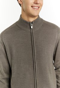 Maglione marrone a costine con zip frontale e colletto alto. Texture a maglia liscia e dettagli con zip argentata nella parte anteriore. Design aderente.